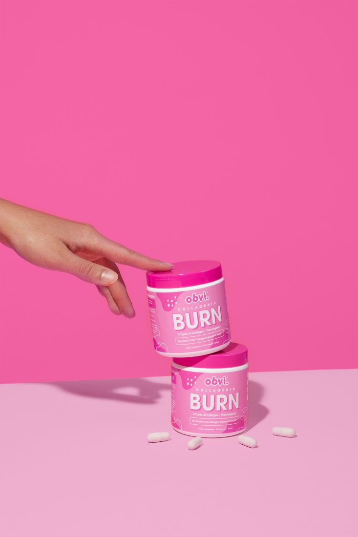 Hand stacking two Collagenic Burn tubs with capsules on a pink surface